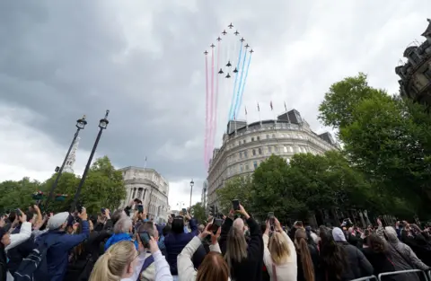 PA Media The Red Arrows and four other military aircraft fly over Trafalgar Square as spectators hold up their phones to photograph the aerial display happening in a cloudy sky.