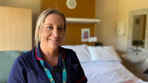Anna Saunders has blonde hair and smiles at the camera in her blue uniform, with a 'cuddlebed' behind her. 