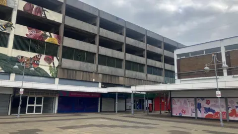 Eccles Shopping Centre multi-story car park. Below the car park there are empty shops, or shops with the shutters down in the middle of the day. The precinct is empty with nobody walking through.