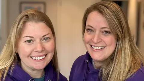 Kate Cooper and Elle Wilks, both wearing branded purple hoodies, smile into the camera.