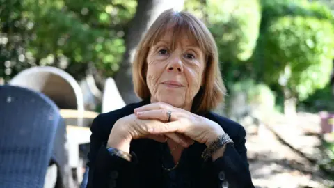 Getty Images A woman with short brown hair wearing a dark blue jumper sits in a sunlit garden. Gisèle Pelicot's hands are crossed under her chin and she looks into the camera