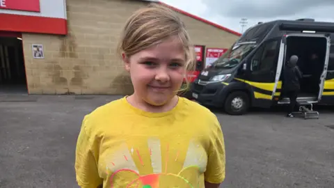 Mia wearing a yellow t-shirt with an image of Pudsey on it. She has her hair tied up and her hands behind her back smiling at the camera. She is standing outside and there is a black van behind her with people in the doorway of it.