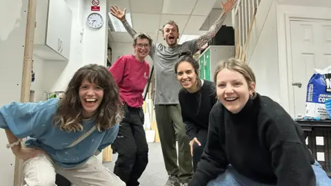 A group of four women and one man smile straight at the camera. Three of the women are squatting down in the foreground. The man at the back at his arms lifted into the air. They are in a white office-type room.