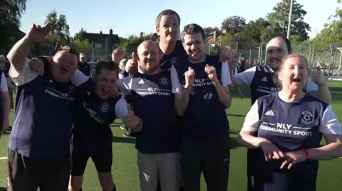A group of people wearing matching “NLY Community Sport” jerseys stand together on an outdoor sports pitch, some raising thumbs and arms toward the camera.

The scene takes place on a sunny day with a fenced field, trees, and buildings visible in the background.

The group is gathered closely in the foreground, suggesting a team or community activity moment.