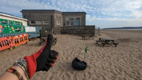  Sophie's super litter picking Ms Newton holding a thumbs up in front of the camera, wearing a black and red glove. Behind her hand there is a patch of sand which she has cleaned up, with a bin bag and litter picking tool visible