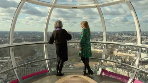 Arsitek Julia Barfield dan reporter BBC Wendy Hurrell berbicara bersama di dalam kapsul di mata London, pemandangan langit dan penjaga kuda di belakang mereka