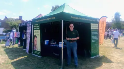 John Copsey John Copsey stands beneath a dark green tent on which the words "Cotswold Prostate Cancer Support Group" appear in yellow. Mr. Copsey wears a dark coloured polo shirt and blue jeans. There are banners containing information n Prostate Cancer to his right and left.