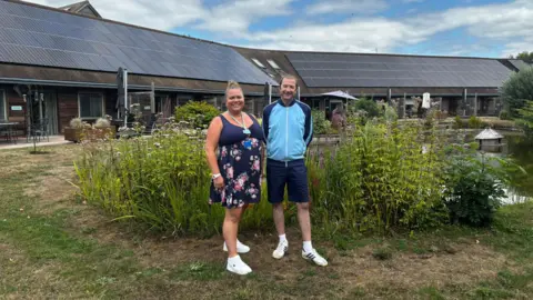 St Richard's Hospice Mark Payne in the hospice gardens with Ria Simons, corporate fundraising lead at St Richard’s Hospice. Mark is wearing a blue tracksuit top, navy blue shorts and white trainers and socks. Ria is wearing a navy blue floral print dress. The two of them are stood in front of a large pond.