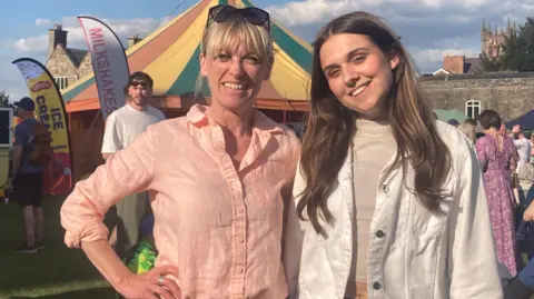 University of Bristol A woman wearing a peach shirt stands with her arm on her hip. A woman wearing a white denim jacket and top stands next to her. A large tent and food signs are behind them.
