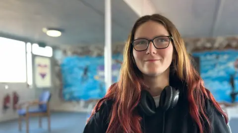 Jon Wright/BBC A young women, with headphones round her neck, sets and smiles at the camera