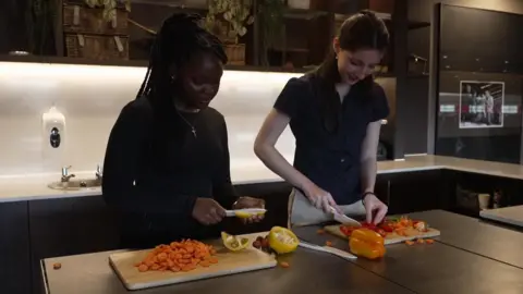 A teenage girl with shoulder-length dark hair wearing a black-top with a crucifix necklace and another girl with shoulder-length dark hair wearing a black short-sleeve shirt and white trousers, chopping vegetables on a kitchen work surface
