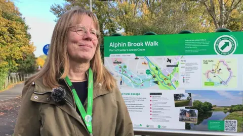 A women wearing an olive green jacket , with glasses and blonde hair, stood in front of a sign, with maps and a description of part of Exeter's green circle.