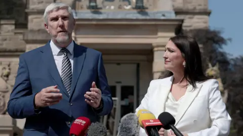 PA Media Emma et John se tiennent à l'extérieur du Stormont ensoleillé devant le groupe de Mike. Emma a de longs cheveux brun foncé, portant un haut avec une veste blanche et un pantalon noir, tandis que John a des cheveux courts gris et une barbe de chèvre gris / blanc, portant un costume bleu marine avec une chemise blanche et une cravate à rayures en diagonale en noir et blanc.