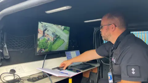 Photograph of Inspector Dan Pickavance. He is pictured inside a police car, with a screen showing images from a drone in the sky. 