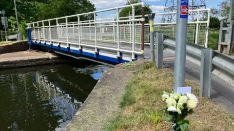 Photo taken from carpark near to the canal. A bridge with a silver car driving on top of it is seen stretching along the width of the canal. 