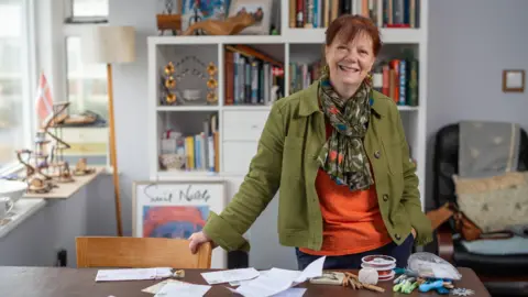 A woman with red hair, wears a green jacket and orange top with a scarf. In front of her is a wooden table with letters strewn across it out of their envelopes.