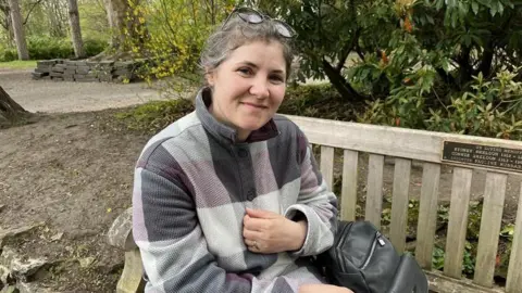 A woman with grey hair is sitting on a bench and holding onto her coat. She is smiling and has sunglasses on her head.