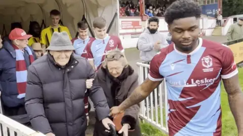 Harry, an elderly man wearing a grey trilby hat and a dark grey puffer coat, shakes the hand of Hastings United player Dylan Pauw as the team leaves the tunnel onto the pitch. Fans can be seen in the stands behind them.