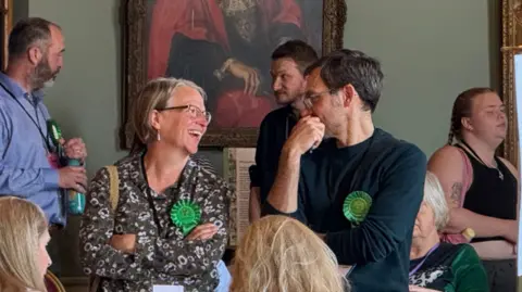 A man and a woman look at each other surrounded by other people. The woman is smiling wearing a dark brown top with a white pattern on it. The man has his right arm and hand to his face. Both have glasses on, the man has a dark top on. Both have green rosettes.