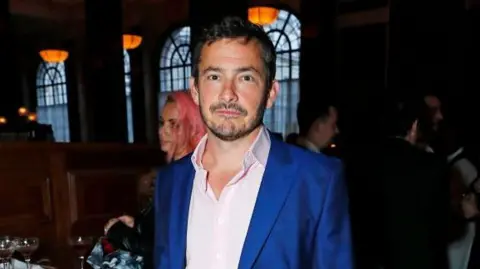 Giles Coren attends the press night performance of "Opening Night" at The Gielgud Theatre on March 26, 2024 in London, England. He wears a blue suit jacket and a pink shirt. It is a head and shoulders shot.