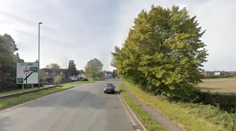 Google The road at the top of a hill. There is one car travelling towards the camera. There is a large square, white roadsign to the left, showing the A5 crossing the road ahead. There is a pub/restaurant beyond the sign, and a large tree to the right of the road and pavement.