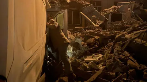 It's nighttime. Karol Swiacki has his back to the camera and he looks through the driver's door of his white transit van, which has been hit by the missile strike. All around him is rubble and collapsed parts of the hotel building. He is using his torch to look in the van and wears jeans, boots, a beige coat and cap.