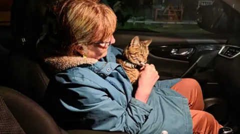 Anne Cornthwaite Anne Cornthwaite sitting in the passenger seat of a car and holding Harvey. She is wearing a blue hooded coat over orange trousers and has Harvey close to her chest while feeding him a treat. It is night time and the picture is taken through the driver's side window.