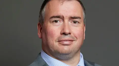 The British parliament is a picture of a smile of Steve Vetheen, an official Parliament of Britain. This is a head picture of a shoulder and wearing a shirt with a gray jacket and blue and white straps