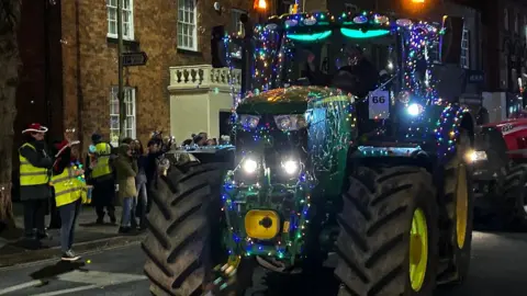 A tractor decorated with fairy lights is driving through the town and people in hi-vis jackets and festive hats are standing nearby.
