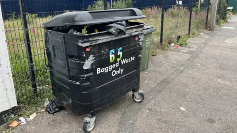 A large black bin labelled "bagged waste only". It's lid is slightly lifted as it's full of waste.