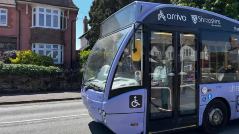 A light purple bus, with 'arriva' and 'Shropshire Council' written on it in white writing. It's driving along a road with a red brick house visible on the other side of the street. The sky is bright blue and there's strong sunshine. 