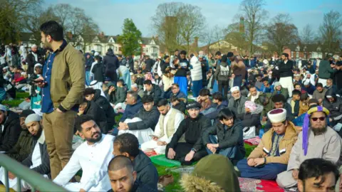 A crowd of men standing or sitting in a park, with trees and houses in the background. Many of them are wearing religious clothing.