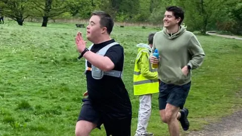 Jillian Perry A man with short hair and black top and shorts and a running hydration vest is running, with another man wearing an olive green hooded top and shorts is smiling while running behind him.