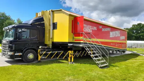 A red and yellow truck with a black cab parked up on a field. On the trailer of the truck is stairs and a ramp. The side of the trailer reads: Travelling Cinema. The Incredible Moving Cinema