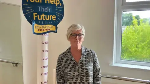 A woman standing next to a sign which looks like a lollipop - it says at the top 'Your Help, Their Future' and a £250,000 target. The pole can be seen from £180,000 and goes up in £10,000 increments until it reaches target. 