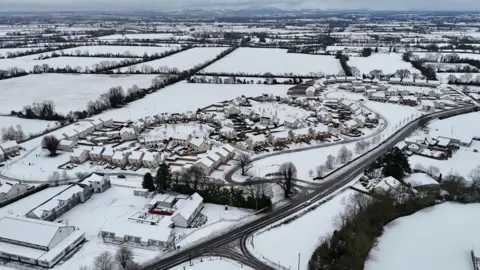 A picture postcard scene of a village in Ireland covered in snow. Roads are visible and some bare trees but everything else, the houses and the fields, are white.