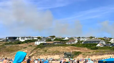 A busy beach is packed with holidaymakers, tents and windbreaks in the foreground, while a row of houses sits on the cliffs above the sand under a bright blue sky. Several grey plumes of smoke can be seen rising behind the homes on the hill.