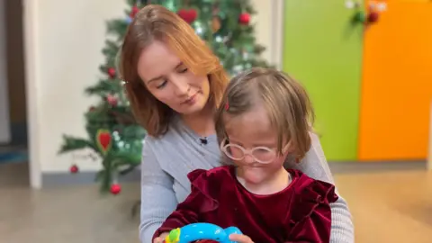 A mother wearing a grey long sleeved top holds her daughter who is wearing glasses and a red velvet dress while she plays with a toy in front of a Christmas tree.