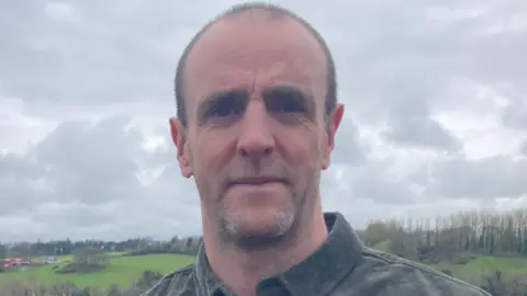 Shows a man in a dark shirt standing in front of fields
