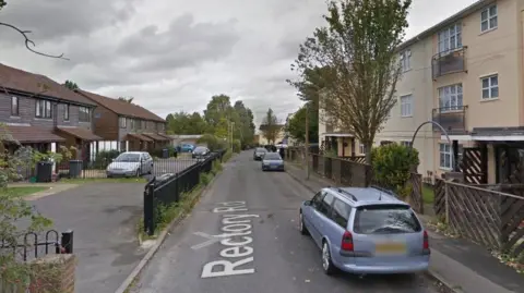 A streetview of Rectory Road on an overcast day. There are a few cars parked along the road, with semi-detached houses on one side and flats on the other. 