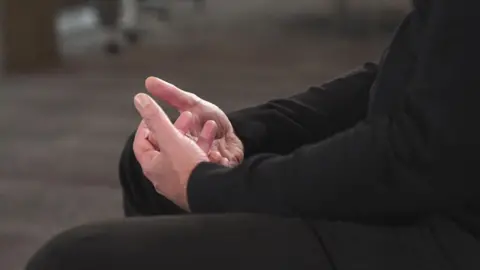 The hands of a man sitting on on a chair, wearing a black jumper and trousers