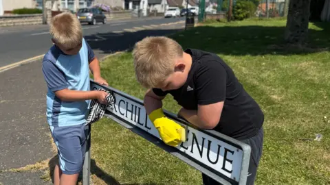 Two children are cleaning a street sign that reads "Churchill Avenue." One child, wearing a light blue shirt, wipes the sign with a checkered cloth, while the other, dressed in a black shirt and wearing yellow rubber gloves, scrubs the sign with a sponge. They are standing beside a grassy verge next to a road. 