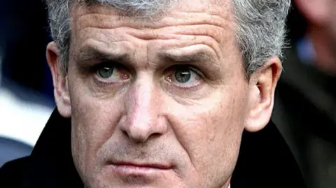 PA Media Image shows Mark Hughes, who has grey hair and is wearing a black overcoat, sitting in the stands of a football stadium