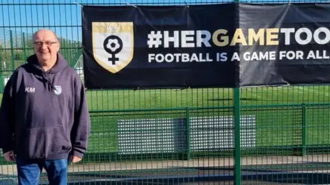 Ken Major A man wearing jeans and a baggy dark hoodie standing next to a green fence with a black Her Game Too sign written in bold white letters on it. There's a football pitch behind the fence.