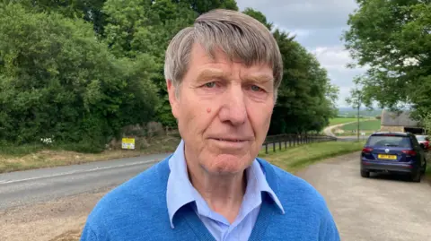 A photo showing a man with grey hair, wearing a blue shirt and blue jumper. He is stood in front of a country road with a car parked in a layby on the right.