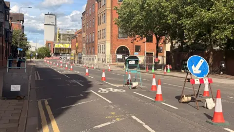 Hull City Council Road works on Alfred Gelder Street. There are rows of orange and white traffic cones in the centre of the road. In the foreground on the right, a “keep left” blue circular road sign with a white arrow is mounted on a small stand, weighted down by sandbags. Metal barriers line the pavement on the right, enclosing a section of the street. Red-brick and light-coloured buildings run along both sides of the road, with trees casting shadows across the scene. In the distance, a bridge-like structure and an industrial-style tower are visible, along with more cones and construction equipment. The sky is partly cloudy.