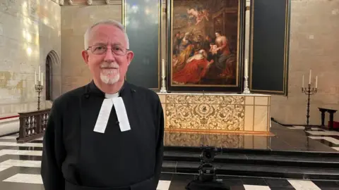 Qays Najm/BBC The Reverend Dr Stephen Cherry in a black cassock, standing in front of a large Flemish Baroque painting.