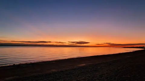 stacey johns A beautiful sunset over a beach. The sky is an orange glow in the distance with the sky turning more blue and dark above. The sea can be seen lit up by the sunset while the beach os covered in darkness.