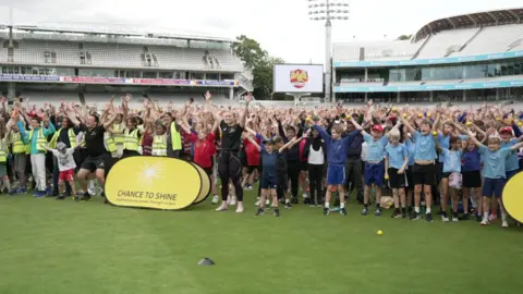 Hundreds of children standing on the pitch at Lord's in a group celebrating, many with their arms in the air. TV Gladiator Fury is in the front row.