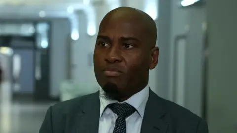 Ike Obi in a dark coloured suit and tie with a white shirt.